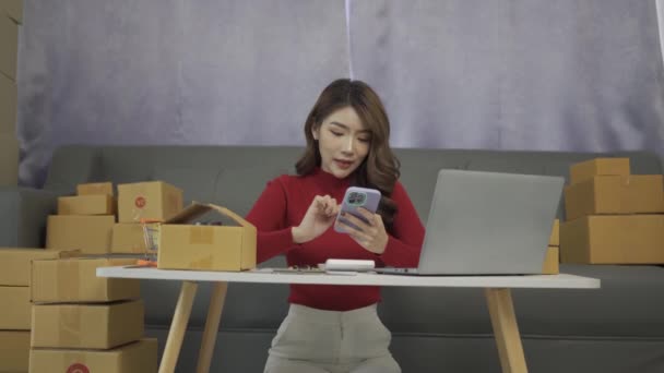 携帯電話を使って購入注文や在庫確認をする若いアジアのビジネス女性小包ボックスノートパソコンで自宅のオフィスで働く中小企業のオーナー オンラインマーケットプレイスの配達 中小企業のビジネスアイデア — ストック動画