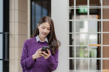 Sosyal medyayı kullanan mutlu genç Asyalı kadın günlük kıyafetler giyen ve akıllı telefonlar kullanan çekici bir kadın internet üzerinden mesajlar gönderiyor ve internette geziniyor..