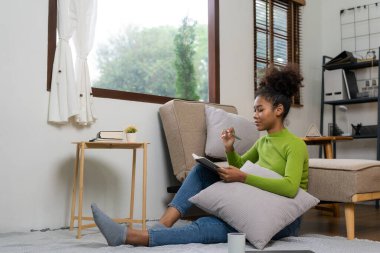 Genç Afrikalı kadın tablet bilgisayar kullanarak oturma odasındaki yumuşak kanepenin yanında mutlu bir şekilde gülümsüyor. Sosyal ağlarda online alışverişin tadını çıkar.