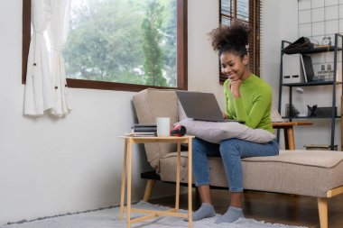 Genç Afrikalı kadın tablet bilgisayar kullanarak oturma odasındaki yumuşak kanepenin yanında mutlu bir şekilde gülümsüyor. Sosyal ağlarda online alışverişin tadını çıkar.