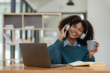 Oturma odasında dizüstü bilgisayarla çalışıp internetten bilgisayar kullanarak öğrenen Afrikalı genç bir üniversite öğrencisi. Gülümseyen kız Webinar ya da uzaktan eğitim dersi izliyor.