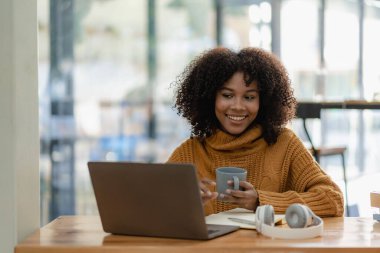 Oturma odasında dizüstü bilgisayarla çalışıp internetten bilgisayar kullanarak öğrenen Afrikalı genç bir üniversite öğrencisi. Gülümseyen kız Webinar ya da uzaktan eğitim dersi izliyor.