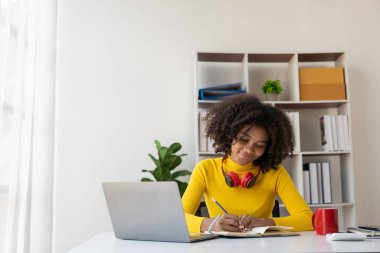 Oturma odasında dizüstü bilgisayarla çalışıp internetten bilgisayar kullanarak öğrenen Afrikalı genç bir üniversite öğrencisi. Gülümseyen kız Webinar ya da uzaktan eğitim dersi izliyor.