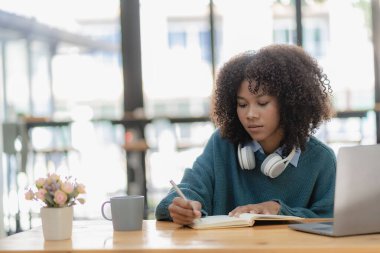 Oturma odasında dizüstü bilgisayarla çalışıp internetten bilgisayar kullanarak öğrenen Afrikalı genç bir üniversite öğrencisi. Gülümseyen kız Webinar ya da uzaktan eğitim dersi izliyor.