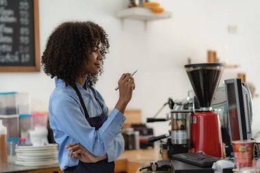 Güzel Afro-Amerikan kadın kasiyer ya da kahveci satın alma detayları yazar ve çekleri hazır eder. Küçük işletme sahibi tezgahın önünde önlük giyen bir kahve dükkanı var..