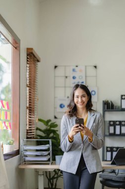 Asyalı iş kadını akıllı telefon kullanarak iş adamıyla konuşuyor ve ahşap masa üzerinde matematik yapıyor. Ofiste dizüstü bilgisayar, vergi, muhasebe ve finans kavramlarıyla çalışıyor.
