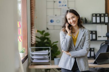 Asyalı iş kadını akıllı telefon kullanarak iş adamıyla konuşuyor ve ahşap masa üzerinde matematik yapıyor. Ofiste dizüstü bilgisayar, vergi, muhasebe ve finans kavramlarıyla çalışıyor.
