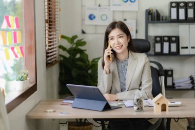 Asyalı iş kadını akıllı telefon kullanarak iş adamıyla konuşuyor ve ahşap masa üzerinde matematik yapıyor. Ofiste dizüstü bilgisayar, vergi, muhasebe ve finans kavramlarıyla çalışıyor.