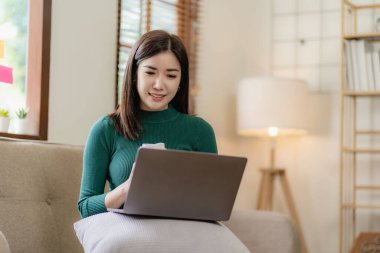 Mutlu Asyalı kız elinde tablet bilgisayar cihazı ile dijital tablet teknolojisini kullanarak evdeki koltukta oturarak internet üzerinden alışveriş yapıyor haberleri okuyor, internette geziniyor, iş buluyor..