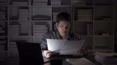 Asyalı erkek işadamı mali belgelerle ve masaüstü bilgisayarlarıyla geç saatlere kadar çalışıyor. Maaşlı çalışan genç adam konsepti 4K