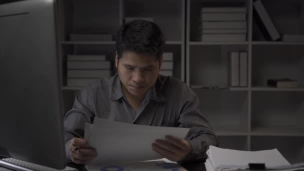 アジアの男性ビジネスマンは 財務文書やデスクトップコンピュータで働いて遅くまでオフィスで働いています 残業サラリーマンコンセプト4K働く若い男 — ストック動画