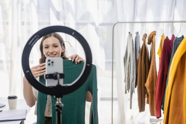 Moda blogcusu konsepti Canlı video, sosyal medya, dijital kamera ile çevrimiçi satış yapmak için kıyafetleri elinde tutan genç kadın..