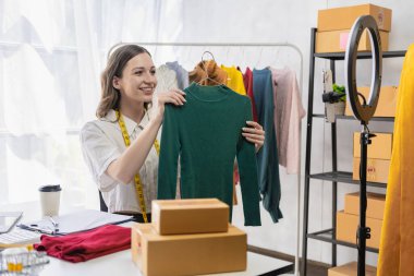 Moda blogcusu konsepti Canlı video, sosyal medya, dijital kamera ile çevrimiçi satış yapmak için kıyafetleri elinde tutan genç kadın..