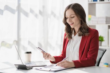 Neşeli bir kadın telefonda kağıt tutarak konuşuyor. Masada oturuyor ve laptop kullanıyor. Modern ofis girişimciliği ve hisse senedi bankacılık sektöründe çalışıyor.