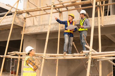 Mimarlar ve mühendisler inşaat planlarını birlikte çiziyor ve inşaat alanında üniforma ve miğfer giyerek büyük sanayi mimarisi hakkında tartışıyorlar..