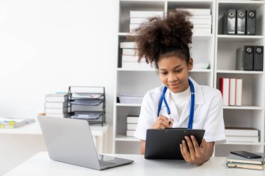 Hastanede çalışan beyaz önlüklü, kulaklıklı bir kadın doktor dizüstü bilgisayarın önünde oturup krampon tutuyor..