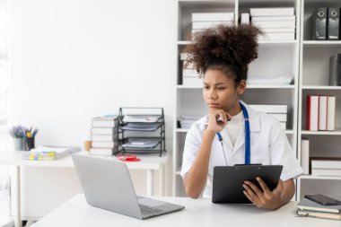 Hastanede çalışan beyaz önlüklü, kulaklıklı bir kadın doktor dizüstü bilgisayarın önünde oturup krampon tutuyor..