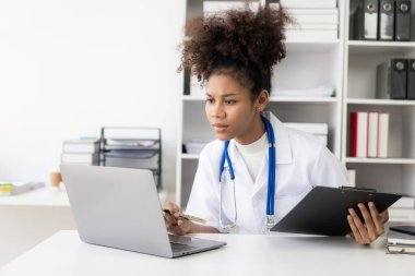 Hastanede çalışan beyaz önlüklü, kulaklıklı bir kadın doktor dizüstü bilgisayarın önünde oturup krampon tutuyor..