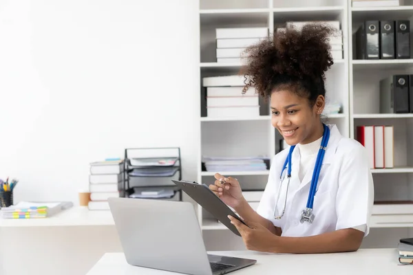 A female doctor working in a hospital in a white coat with headphones sits working in front of a laptop, holding a cleaboard.