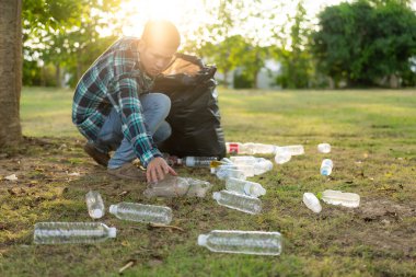 Adamın elleri çöp topluyor, boş plastik su şişeleri, yerde yahni, kuru yapraklar, çöp torbalarında plastik şişeler, temizlik, toprak ekoloji günü konsepti...