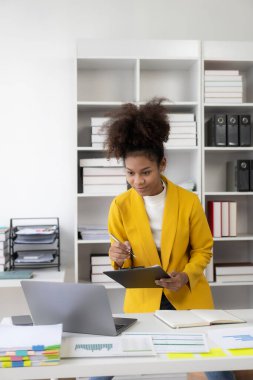 Profesyonel Afro-Amerikan kadın şirket yöneticisi şirket ofisinde takım elbise giyip dizüstü bilgisayar ve finansal muhasebe belgeleriyle çalışıyor. Dikey görünüm