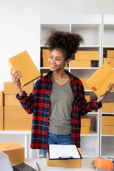 Small business startup SME app African female business owner using laptop receives and verifies online orders to prepare product boxes and ship them. vertical image