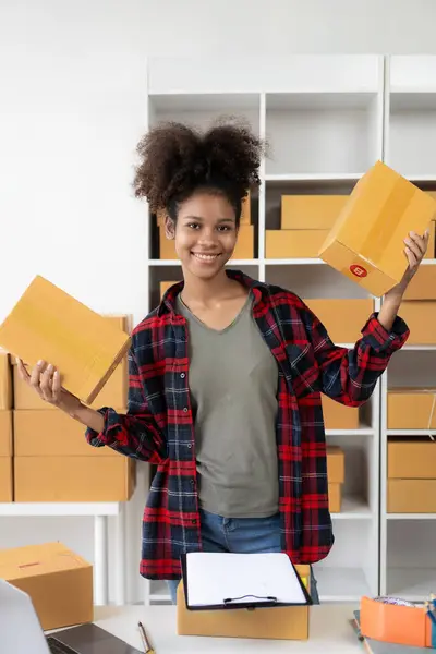 Small business startup SME app African female business owner using laptop receives and verifies online orders to prepare product boxes and ship them. vertical image