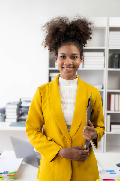Busy professional African American female company manager wearing a business suit in a corporate office working with a laptop computer and financial accounting documents. Vertical view