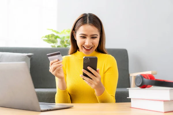 Asian girl pays online using laptop to shop at home Attractive Asian millennial woman holds her smartphone and credit card using mobile banking or online shopping app.