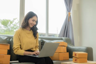 Küçük işletme girişimcisi ya da serbest çalışan Asyalı bir kadın çevrimiçi siparişlerden dizüstü bilgisayarla çalışıyor. Paketleme kutuları, çevrimiçi pazarlama, KOBİ işleri ve nakliye işleri ile başarılı Asyalı genç bir kadın..