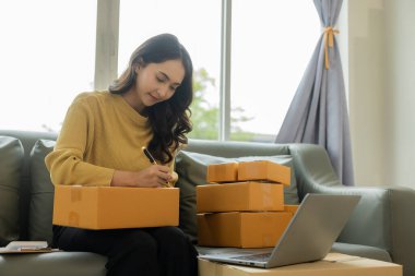 Küçük işletme girişimcisi ya da serbest çalışan Asyalı bir kadın çevrimiçi siparişlerden dizüstü bilgisayarla çalışıyor. Paketleme kutuları, çevrimiçi pazarlama, KOBİ işleri ve nakliye işleri ile başarılı Asyalı genç bir kadın..
