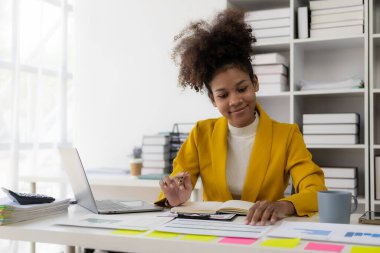 Güzel Afro-Amerikalı iş kadını dizüstü bilgisayar ve finansal belgelerle çalışıyor ofiste evrak yazıyor, e-postaları kontrol ediyor, iş yerinde online toplantıya katılıyor.
