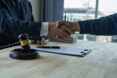 Bir avukat, avukatın bürosundaki müvekkilleriyle belgeler, sözleşmeler, anlaşmalar, iş birliği anlaşmaları yapan bir müvekkille el sıkışır. Masanın üzerinde adaletin çekiciyle.