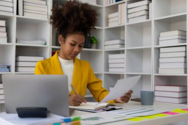 Dizüstü bilgisayarla çalışan Afrikalı Amerikalı iş kadını. Finansal tablolar ve grafikler. Ofisteki kağıt belgelerdeki verileri kontrol ediyor.