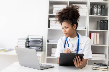 Klinikte çalışan, bilgisayarla klinikte çalışan, laptopa bakan, hastalarla video konferansı yapan, online danışmanlık konsepti olan Afro-Amerikalı genç bir kadın..