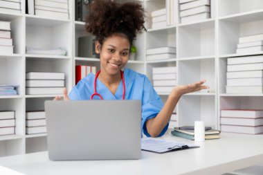 Klinikte çalışan, bilgisayarla klinikte çalışan, laptopa bakan, hastalarla video konferansı yapan, online danışmanlık konsepti olan Afro-Amerikalı genç bir kadın..