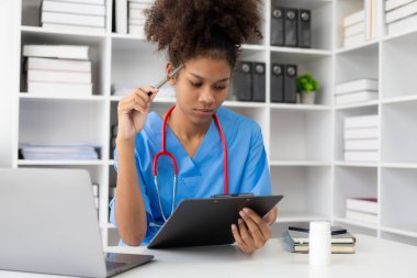 Klinikte çalışan, bilgisayarla klinikte çalışan, laptopa bakan, hastalarla video konferansı yapan, online danışmanlık konsepti olan Afro-Amerikalı genç bir kadın..