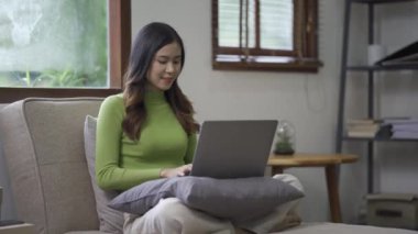 Çekici Asyalı kadın çalışıyor, internette sörf yapıyor, dizüstü bilgisayar kullanıyor, evden oturma odasına geçip neşeli bir ifadeyle çalışıyor..