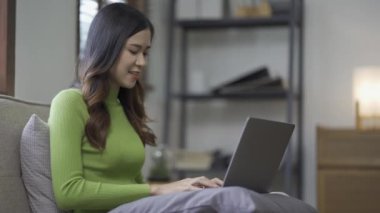 Çekici Asyalı kadın çalışıyor, internette sörf yapıyor, dizüstü bilgisayar kullanıyor, evden oturma odasına geçip neşeli bir ifadeyle çalışıyor..