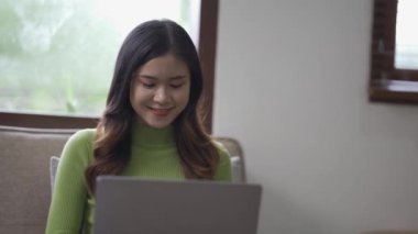 Çekici Asyalı kadın çalışıyor, internette sörf yapıyor, dizüstü bilgisayar kullanıyor, evden oturma odasına geçip neşeli bir ifadeyle çalışıyor..