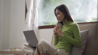 Çekici Asyalı kadın çalışıyor, internette sörf yapıyor, dizüstü bilgisayar kullanıyor, evden oturma odasına geçip neşeli bir ifadeyle çalışıyor..