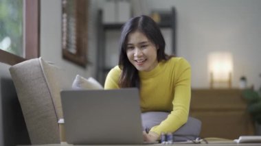 Çekici Asyalı kadın çalışıyor, internette sörf yapıyor, dizüstü bilgisayar kullanıyor, evden oturma odasına geçip neşeli bir ifadeyle çalışıyor..