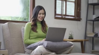 Çekici Asyalı kadın çalışıyor, internette sörf yapıyor, dizüstü bilgisayar kullanıyor, evden oturma odasına geçip neşeli bir ifadeyle çalışıyor..