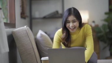 Çekici Asyalı kadın çalışıyor, internette sörf yapıyor, dizüstü bilgisayar kullanıyor, evden oturma odasına geçip neşeli bir ifadeyle çalışıyor..