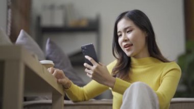 Sosyal medya için akıllı telefon kullanan gülümseyen Asyalı kadın, evde oyunlar oynayan, mutlu hisseden, evde mesajlaşan ve alışveriş yapan. Evdeki yaşam tarzı kadını