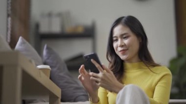 Sosyal medya için akıllı telefon kullanan gülümseyen Asyalı kadın, evde oyunlar oynayan, mutlu hisseden, evde mesajlaşan ve alışveriş yapan. Evdeki yaşam tarzı kadını