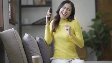 Sosyal medya için akıllı telefon kullanan gülümseyen Asyalı kadın, evde oyunlar oynayan, mutlu hisseden, evde mesajlaşan ve alışveriş yapan. Evdeki yaşam tarzı kadını