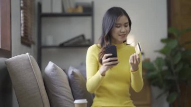Kanepede otururken, elinde online alışveriş için kredi kartı tutan Asyalı bir kadın akıllı telefon kullanıyor ve evde internet üzerinden alışveriş yapıyor.