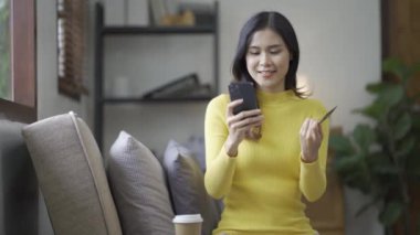 Kanepede otururken, elinde online alışveriş için kredi kartı tutan Asyalı bir kadın akıllı telefon kullanıyor ve evde internet üzerinden alışveriş yapıyor.