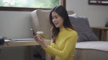 Kanepede otururken, elinde online alışveriş için kredi kartı tutan Asyalı bir kadın akıllı telefon kullanıyor ve evde internet üzerinden alışveriş yapıyor.